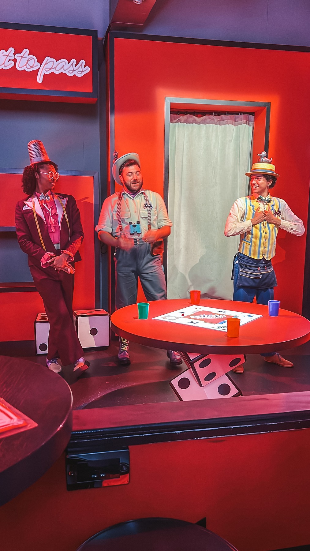 Three Monopoly Lifesized hosts in colourful costumes around a red table with giant dice and a Monopoly board